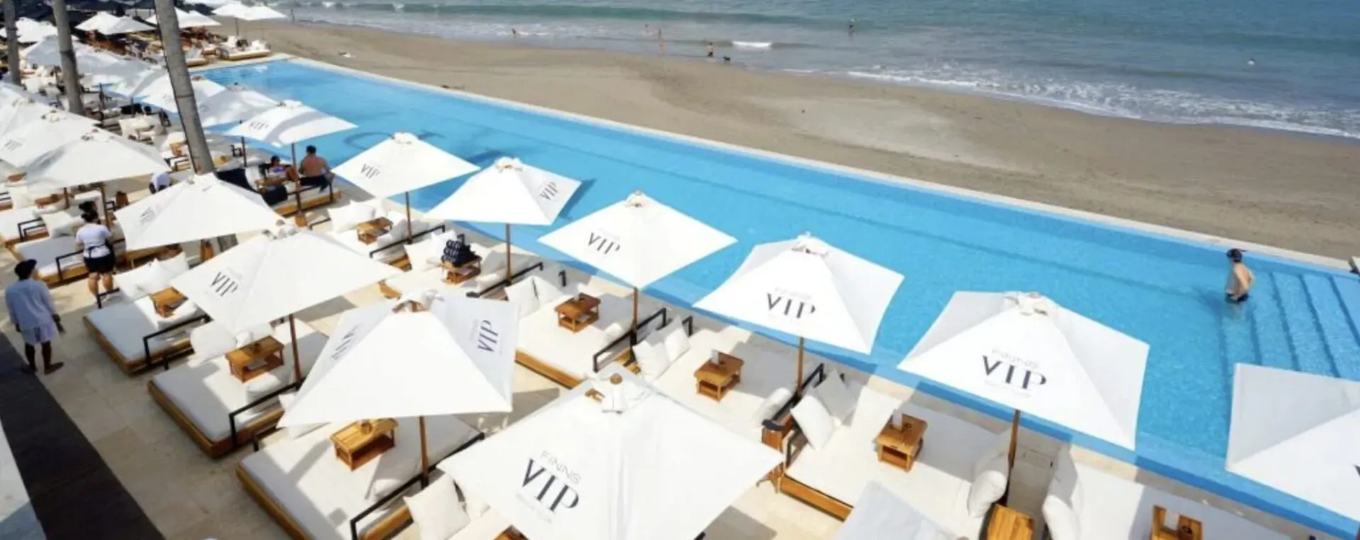 Aerial view of luxurious beachside infinity pool with white VIP umbrellas over loungers, sandy beach, and ocean.