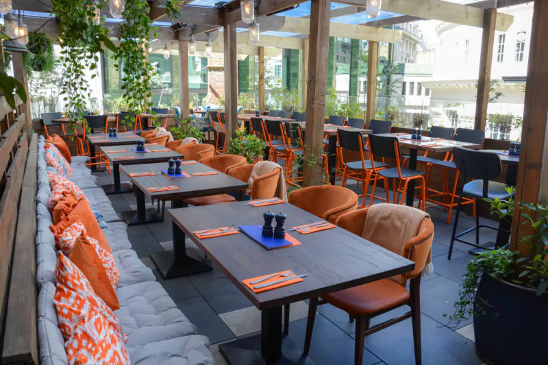 CoqBull Soho outdoor terrace with orange chairs, cushioned benches, wooden tables set with blue pepper shakers, plants, and lanterns.