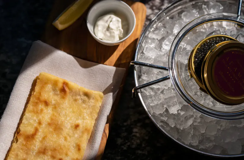 Caviar in ice bucket on silver tray with blini, crème fraîche, and lemon wedge on wooden board