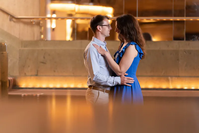 Couple in white shirt and blue dress embrace intimately in Sheraton Grand London Park Lane gallery, warm lighting.