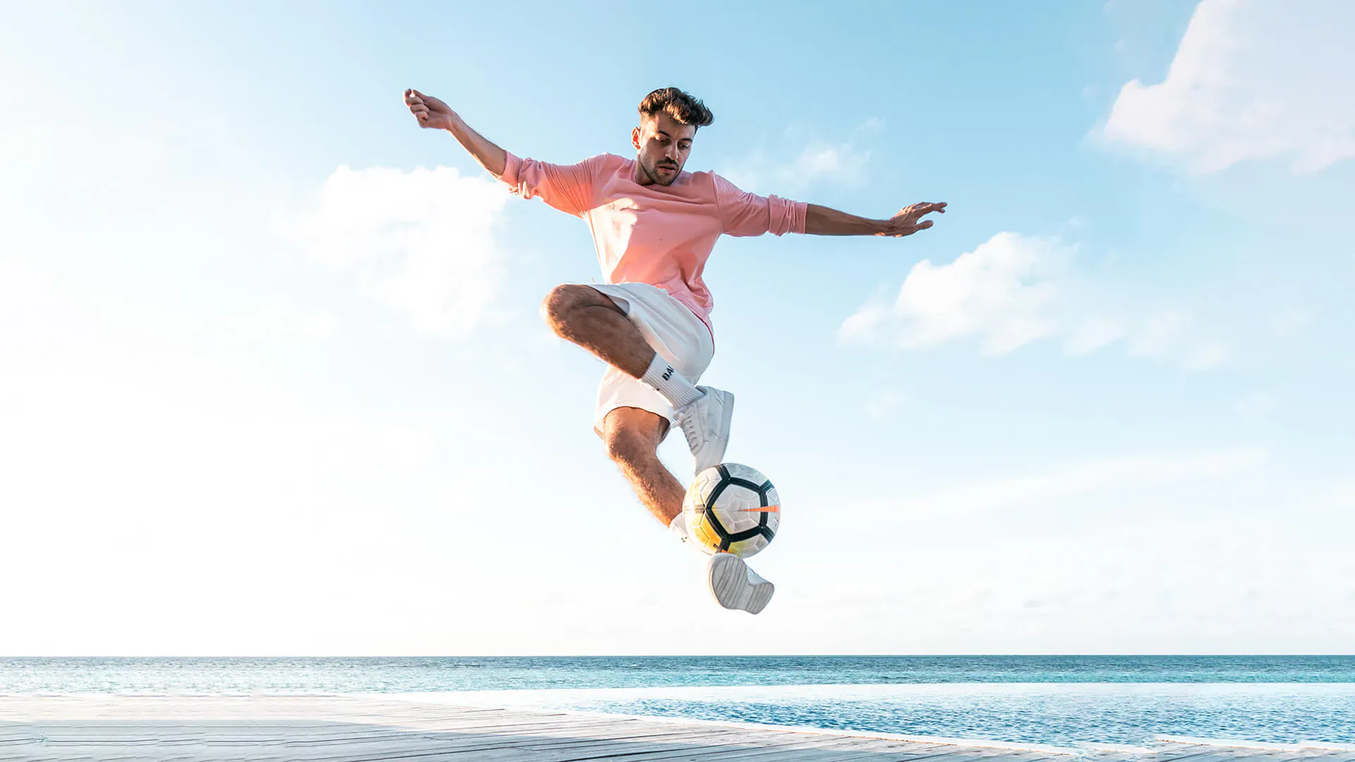Man in pink shirt and white shorts jumps kicking soccer ball on beach at Lily Beach Resort, sunny sky
