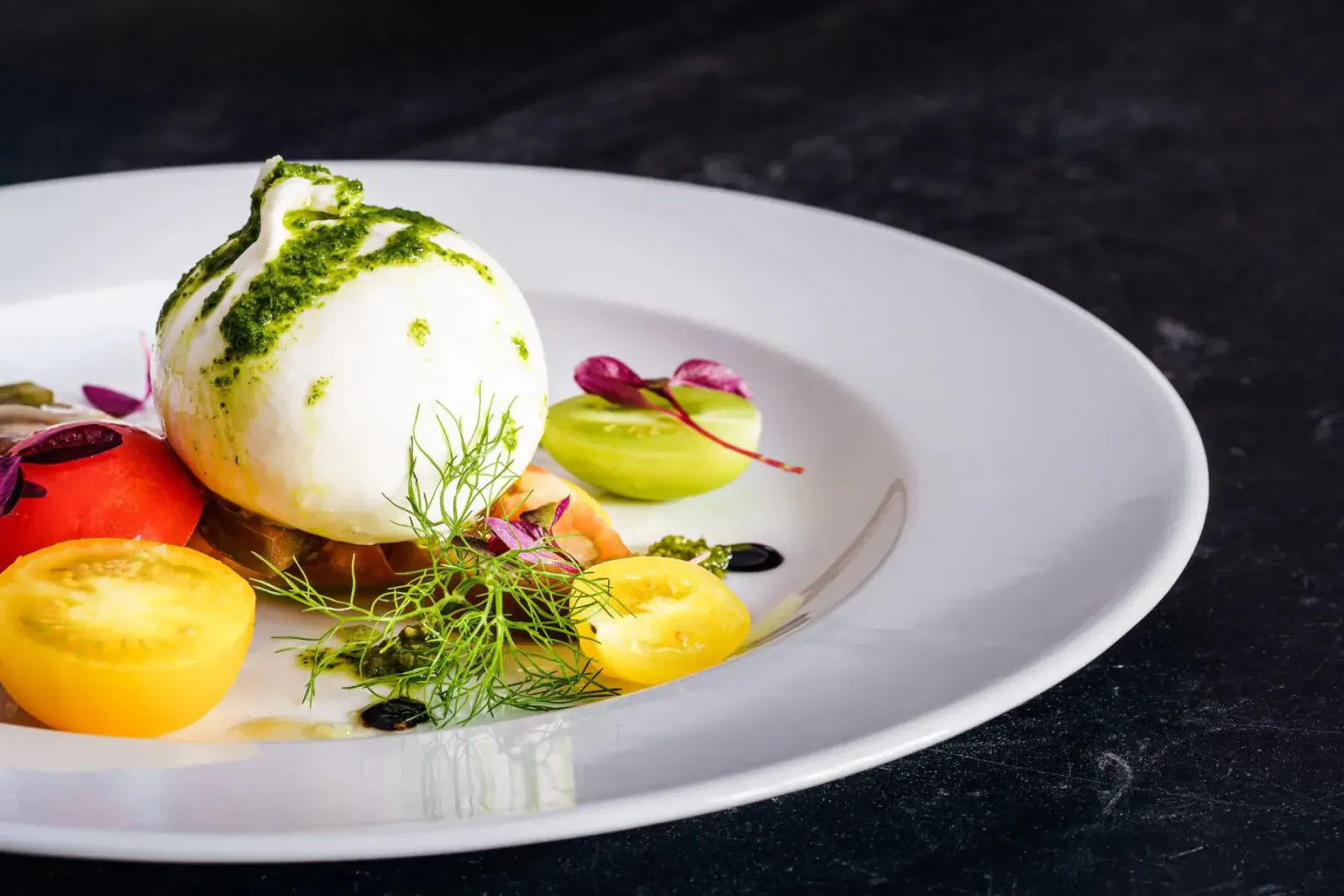 White burrata with green pesto, yellow tomatoes, pink radish, dill on white plate