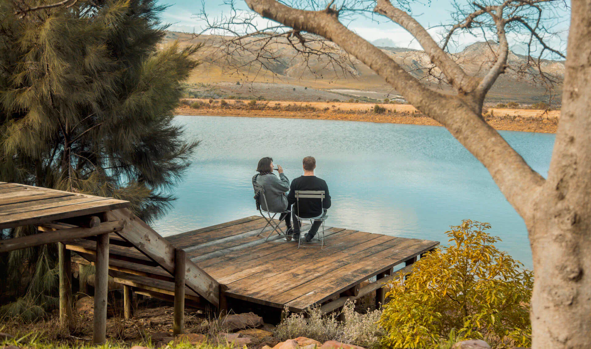 Couple seated on chairs on a wooden dock overlooking a serene lake at South Hill Vineyards, with trees and mountains.