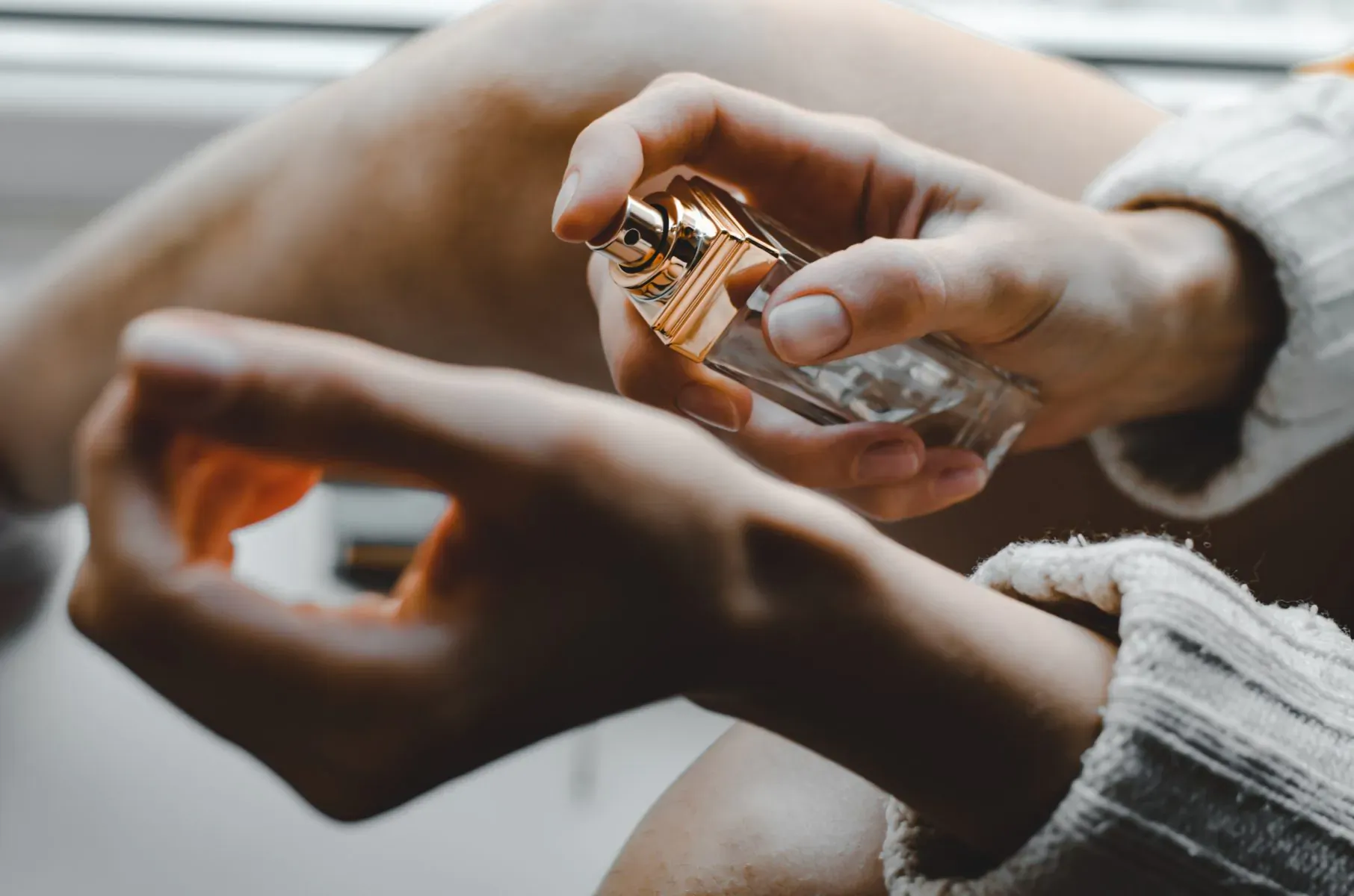 Close-up of hands in cozy sweater holding and spraying gold-capped perfume bottle by window