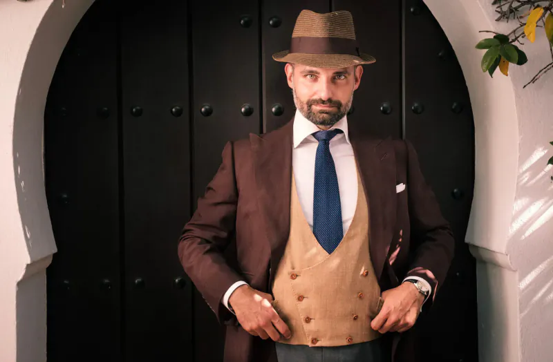Man in brown fedora, suit, blue tie, and beige waistcoat stands confidently before arched wooden door in Marbella courtyard.