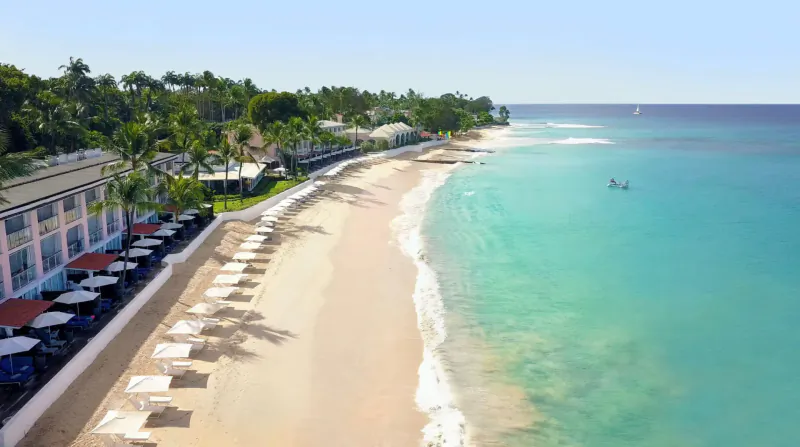 Aerial view of Fairmont Royal Pavilion resort along a sandy beach with turquoise ocean, palm trees, and umbrellas.