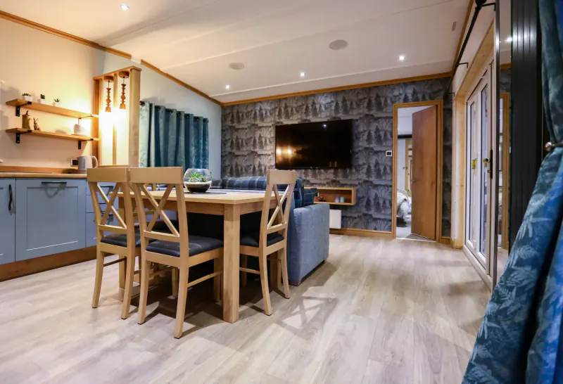 Spacious holiday lodge interior with wooden dining table, blue kitchen, blue sofa, TV, and nautical wallpaper.