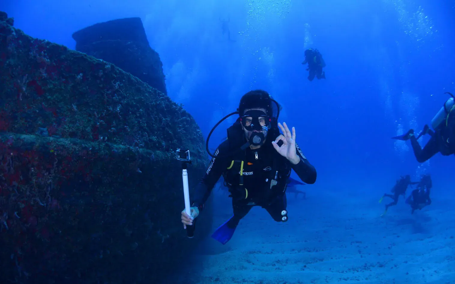 Diver making peace sign with GoPro while swimming near coral reef with other divers underwater