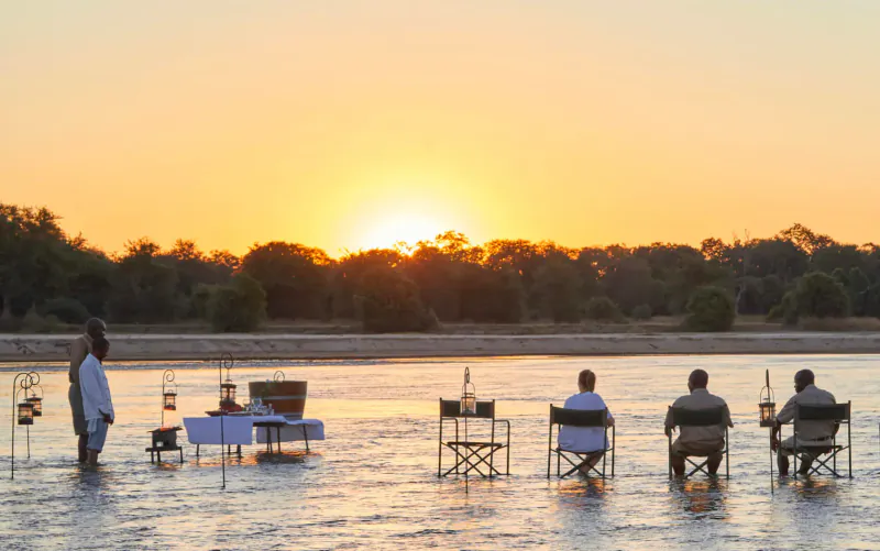 Sunset riverside dinner setup at Mfuwe Lodge with lanterns, table, chairs, and distant trees.