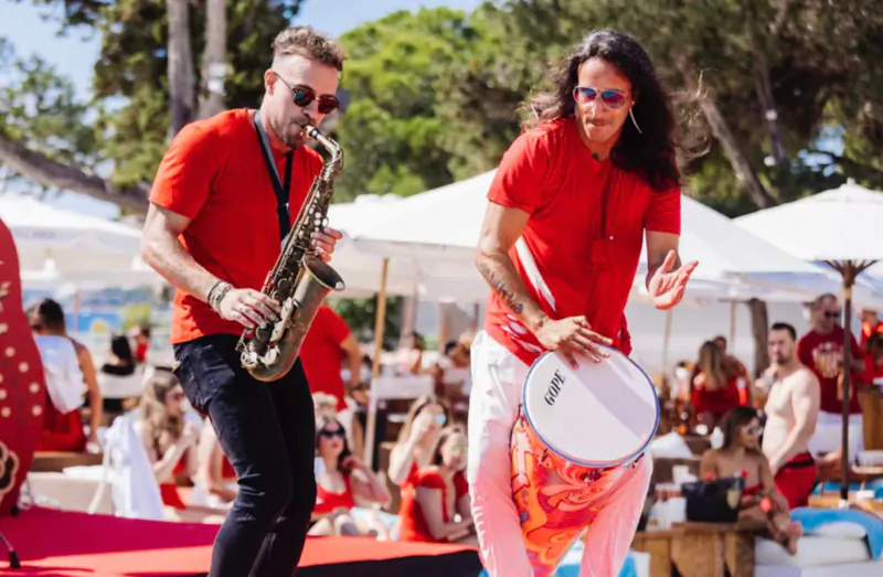 Man playing red saxophone and woman with white tambourine performing energetically on Nikki Beach Ibiza stage amid beach crowd.