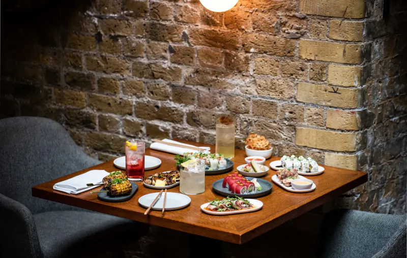 Assortment of sushi, sashimi, cocktails, and small plates on wooden table against brick wall in cozy tavern setting.
