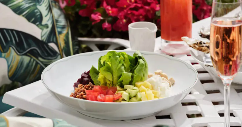 Vibrant salad with lettuce, tomatoes, cucumber, feta in white bowl on outdoor table with pink flowers, rosé glass, red drink