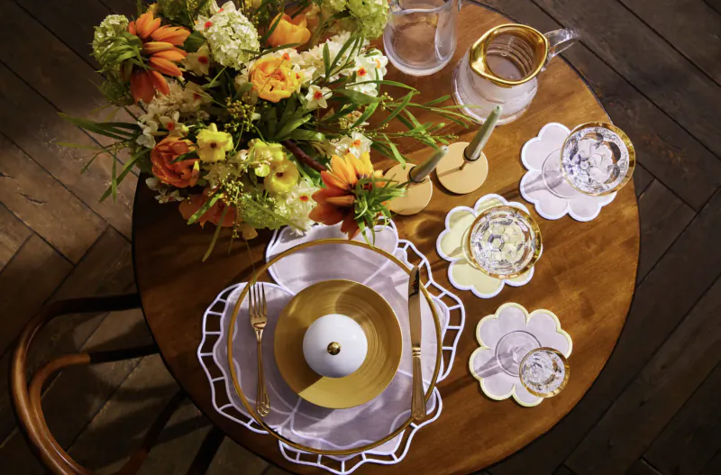 Luxury table setting with vibrant orange and yellow flower bouquet, gold pitcher, white plates, and lace doilies on wooden table.