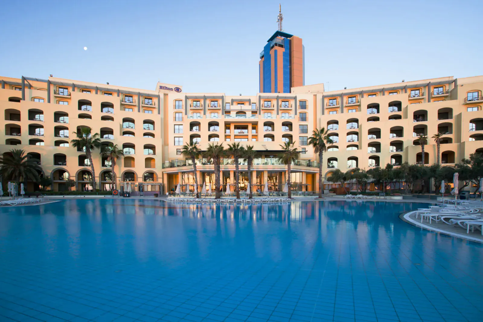 Hilton Malta hotel with tall tower, arched facade, palm trees, and large blue pool at sunset.