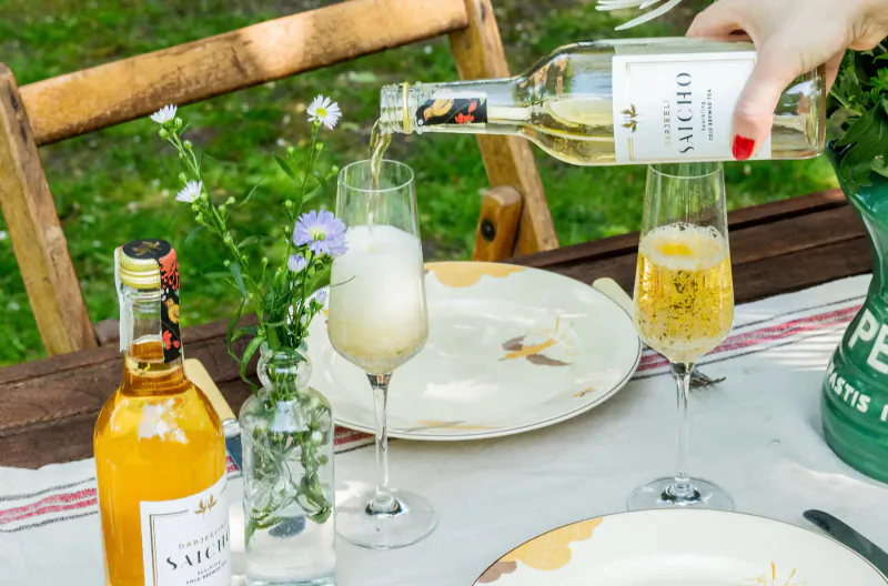 Hand pouring Saicho sparkling tea from bottle into flute glass on outdoor wooden table with wildflowers.