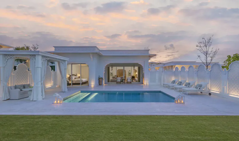 Luxury white villa with arched pergola, lounge seating, lit pool, and lawn at sunset in Qatar.