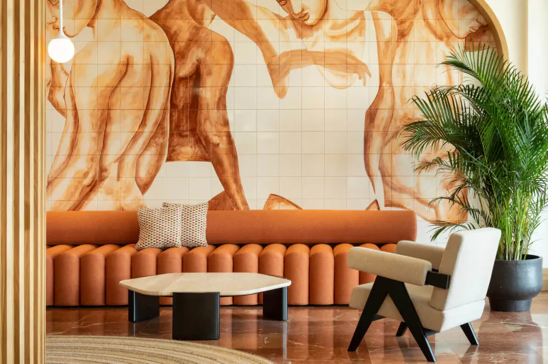 Hotel Riomar lobby: arched orange abstract nude figures mural, orange tufted sofa, black table, white chair, palm plant.