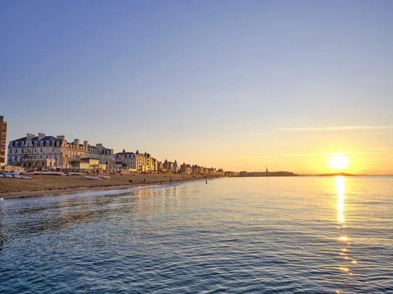 Saint Malo, France: luxury buildings line sandy beach at sunset, calm sea reflecting golden sun.