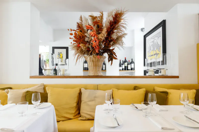 Cozy restaurant booth with yellow cushions, white table set with wine glasses, large pampas flower arrangement, wine bottles on wooden shelf behind.