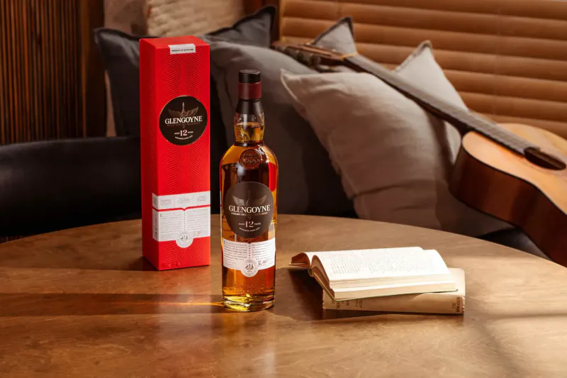 Tamdhu single malt whisky bottle and red box on wooden table with open book, pillows, and guitar