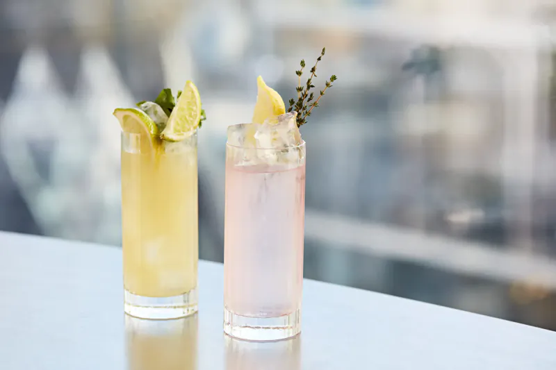 Two tall cocktails on a bar counter: yellow with lime and herbs, pink with lemon, city skyline view beyond.