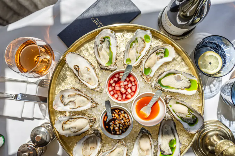 Elegant overhead view of fresh oysters on ice in gold tray with sauces, rosé, champagne at fine dining table.