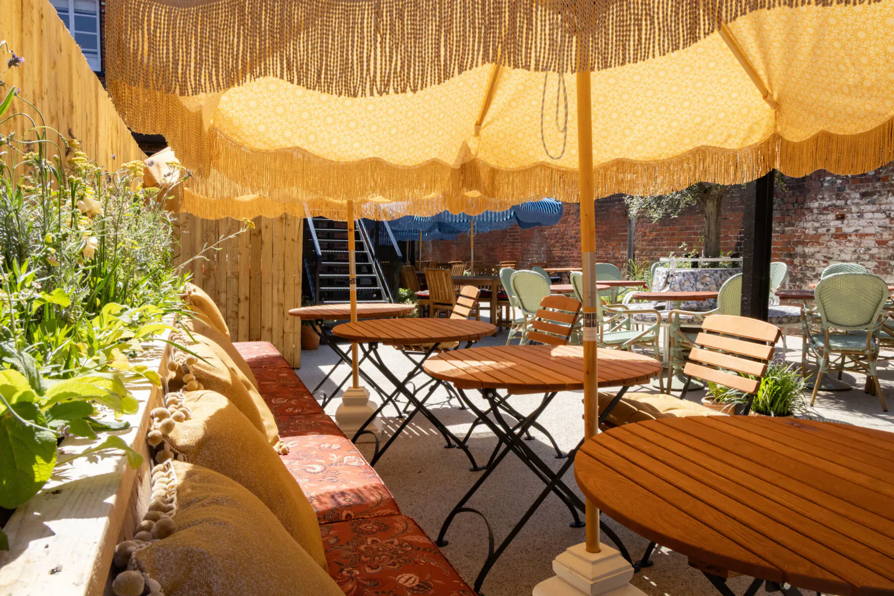 Outdoor seating at The George & Dragon Marlow: fringed yellow umbrellas, wooden tables, green chairs, plants, brick wall, stairs.