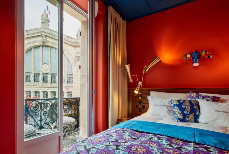 Cozy red hotel room with blue ceiling, colorful bed, arched lamp, balcony view of grand Paris train station.