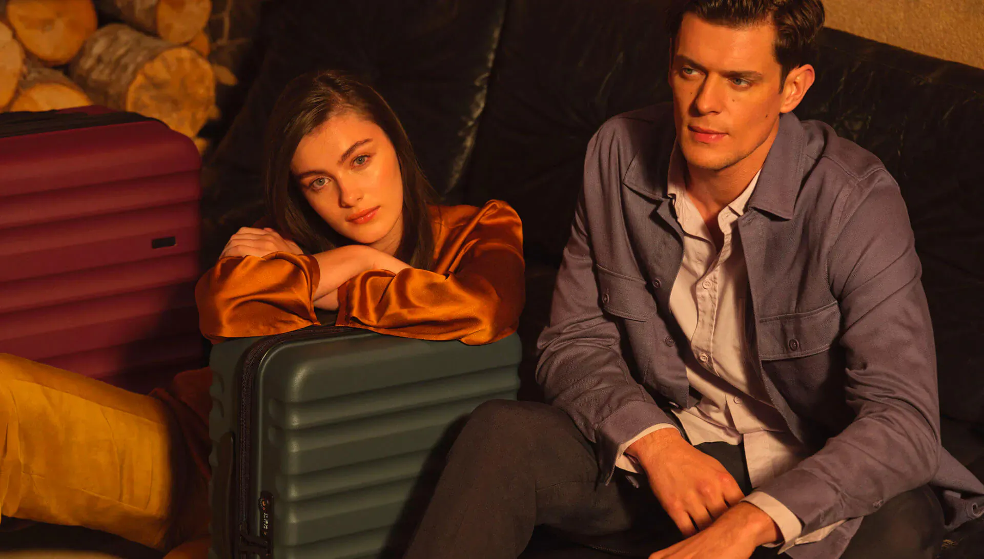 Woman in orange velvet and man in blue blazer lounging on velvet sofa with green and purple Antler Clifton suitcases beside firewood.