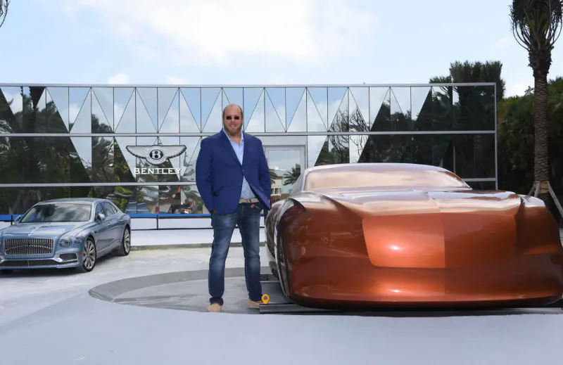 Bald man in blue blazer and sunglasses stands beside sleek copper Bentley concept car at Bentley showroom with palms and glass facade