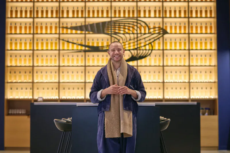 Smiling man in navy robe and beige scarf stands at L’Indigo bar counter, backlit by glowing cognac bottles and Martell bird logo.