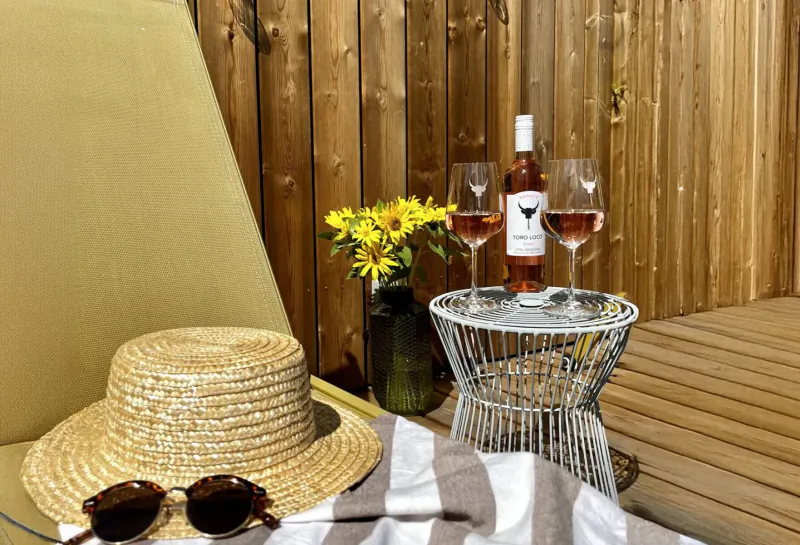 Two glasses of rosé wine on a table with bottle, sunflowers, straw hat, sunglasses, towel by wooden fence