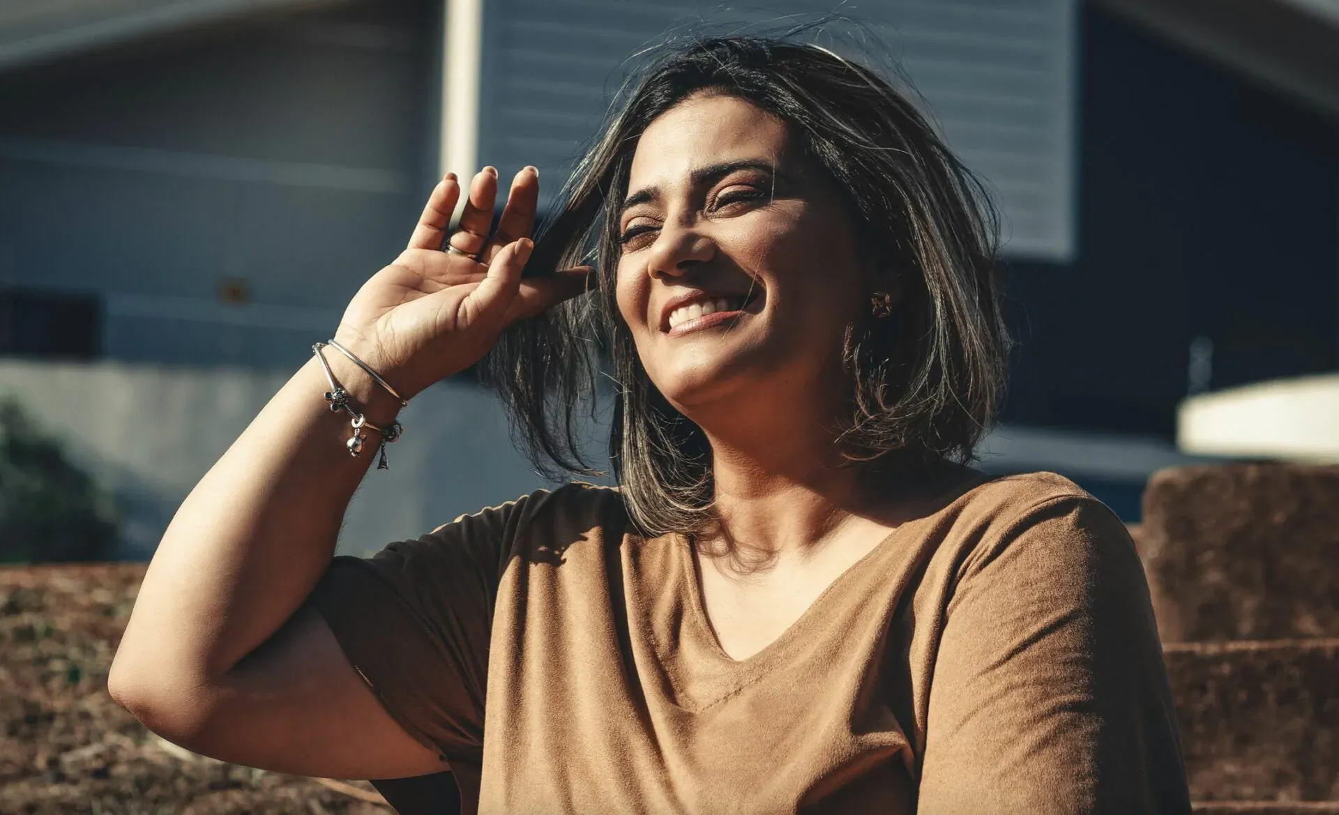 Smiling woman with shoulder-length dark hair raises hand to face, wearing beige V-neck top outdoors against brick wall