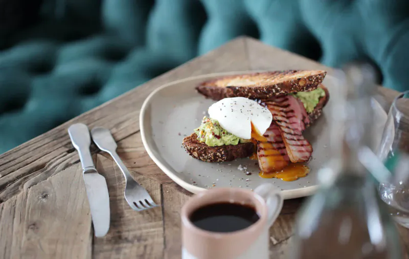 Poached eggs on seeded toast with avocado and bacon on a plate, beside coffee cup on rustic wooden table against velvet booth.