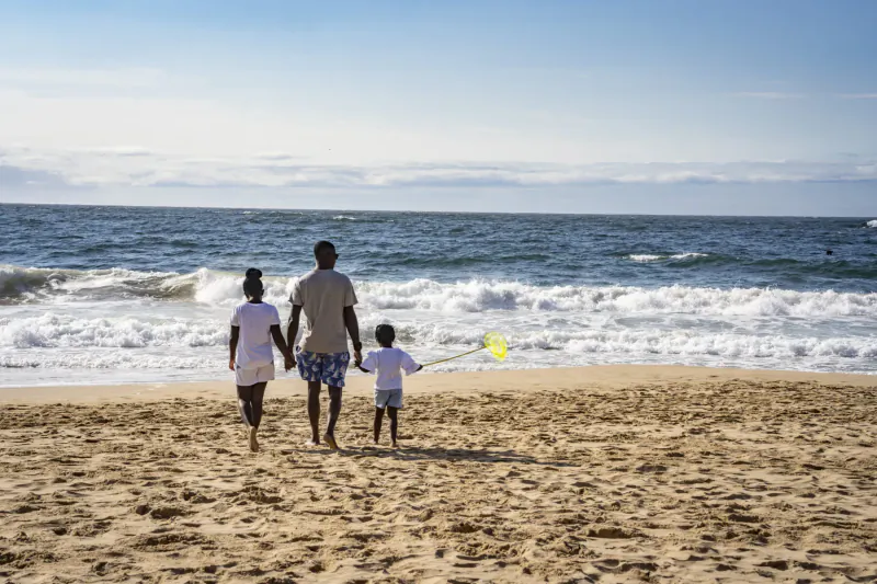 Black family walking on sunny beach: dad holding kids' hands, boy with yellow beach paddle, ocean waves ahead