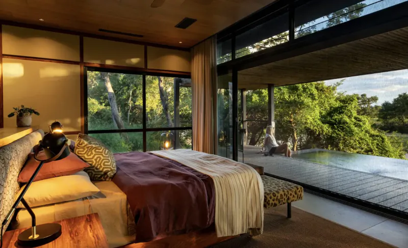 Luxurious wooden bedroom in Tanda Tula lodge with large windows overlooking lush green trees, infinity pool, and distant figure.