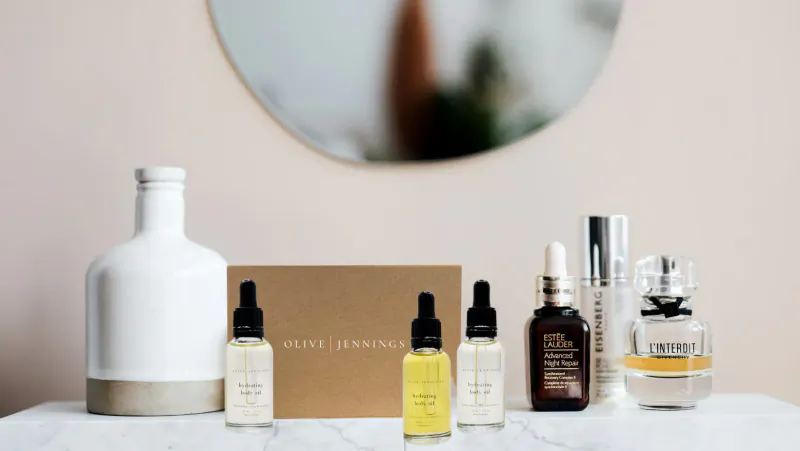 Assortment of Olive Jennings skincare: white vase, serum dropper bottles, gold box, and perfume on marble table with round mirror.