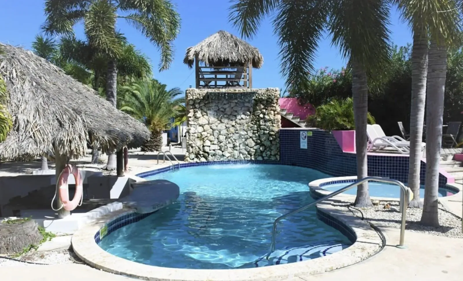 Tropical Bonaire resort pool with blue water, hot tub, stone waterfall, thatched huts, palms, and lounge chairs