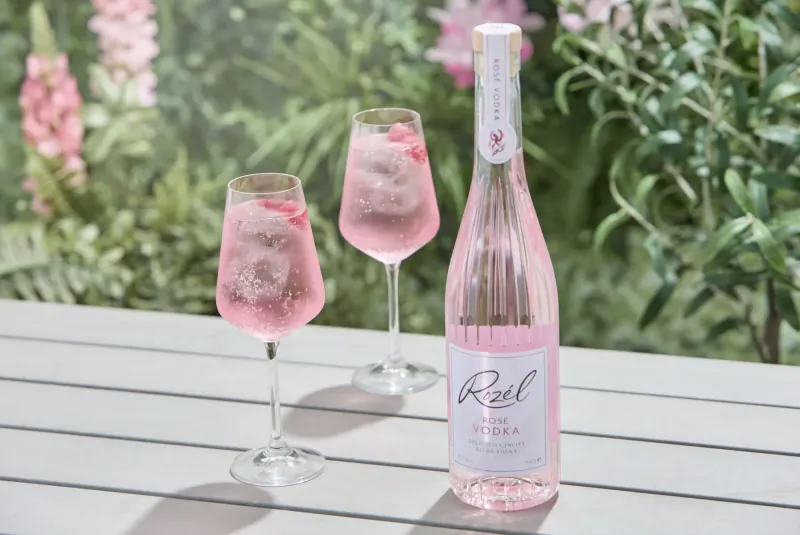 Rêve Rosé bottle and two pink fizzing glasses on a table amid garden greenery.