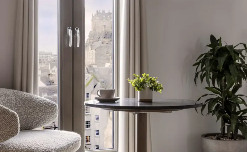 Cozy hotel room corner with curved chair, small round table holding white coffee cup and yellow flowers, tall plant, open doors to Edinburgh Castle view.