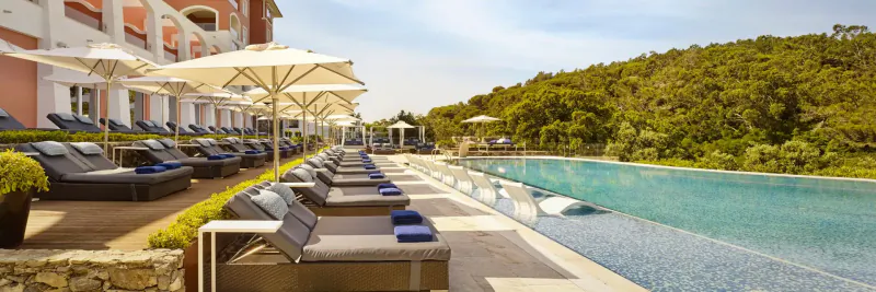 Luxury infinity pool at Penha Longa Resort, Portugal, with loungers, umbrellas, towels, and green hills backdrop.