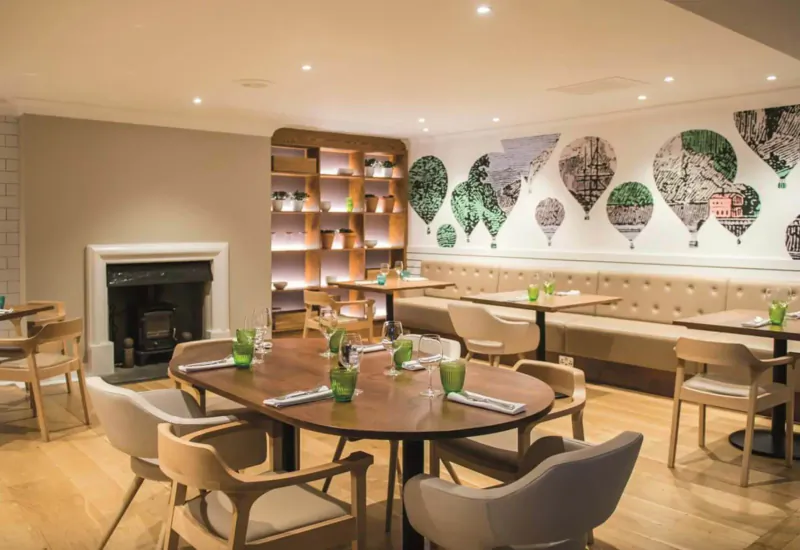 Modern hotel dining area with oval wooden table, green glasses, beige banquette, hot air balloon wall mural, and fireplace.