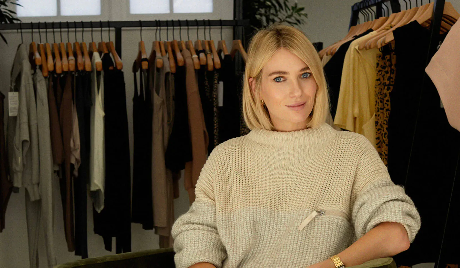Blonde woman in cream sweater smiles at camera in Varley clothing store with racks of dresses behind her