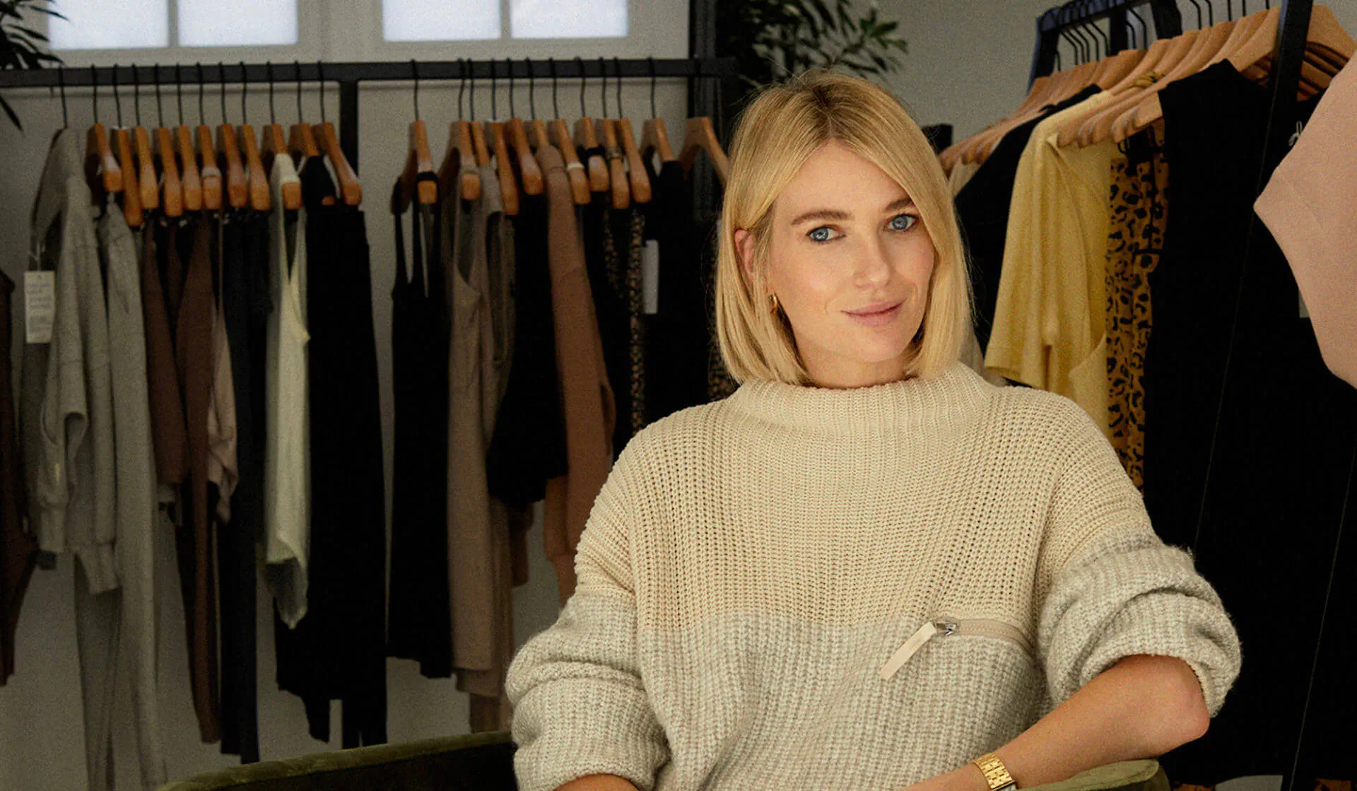 Blonde woman in cream sweater smiles at camera in Varley clothing store with racks of dresses behind her
