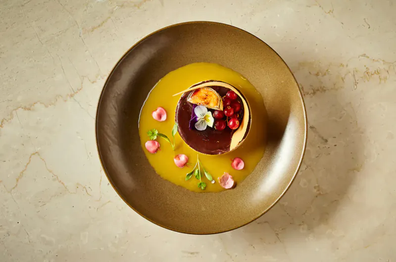 Top-down view of panna cotta dessert in brown bowl with orange sauce, orange segment, red berries, and edible flowers on marble surface