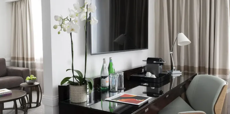 Elegant hotel room desk with coffee maker, green water bottles, orchids, TV, lamp, and sofa in luxurious setting.