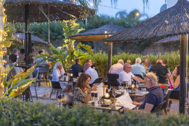 Diverse group dining at outdoor tropical restaurant at dusk, under thatched umbrellas with string lights and palms.