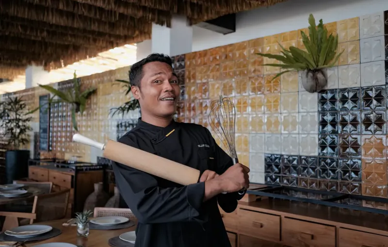 Smiling chef in black uniform holding large rolling pin and skewers, in tropical tiled seaside dining area