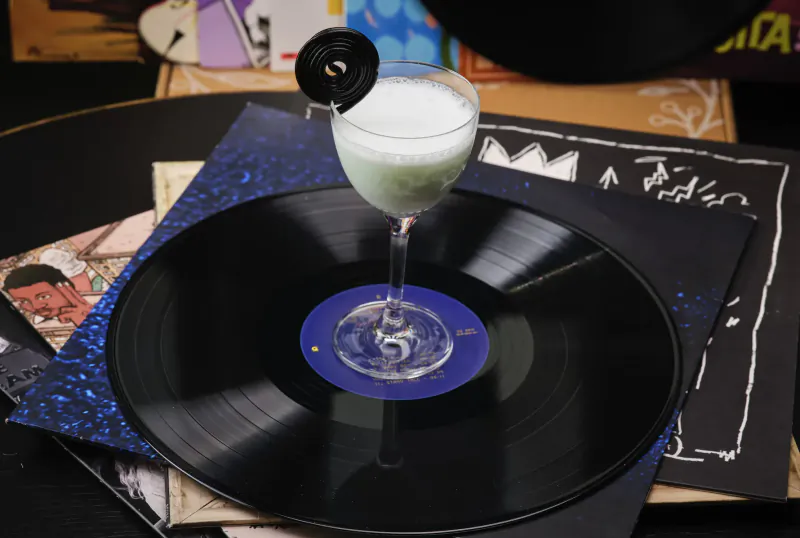 Milky cocktail in martini glass on vinyl record with Jean-Michel Basquiat crown art, surrounded by records at rooftop bar.