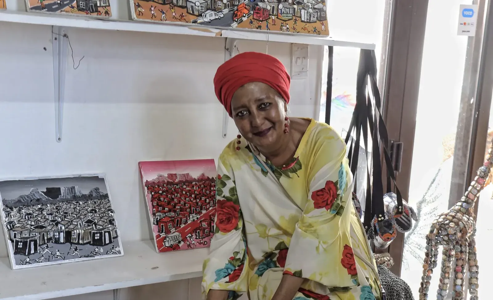 Smiling Cape Malay woman in red headscarf and yellow floral dress sits by art shelves and giraffe statue in shop.
